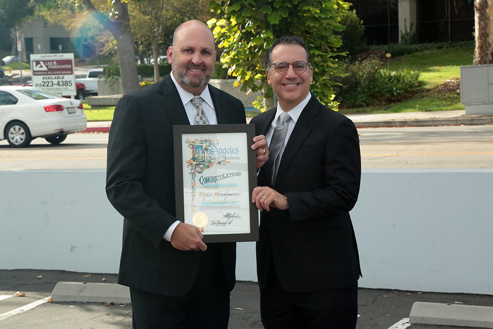 Two men in suits stand outdoors in a parking lot, smiling at the camera during a CHQS 20th anniversary celebration. One holds a framed certificate that reads Congratulations. Trees and buildings are visible in the background.