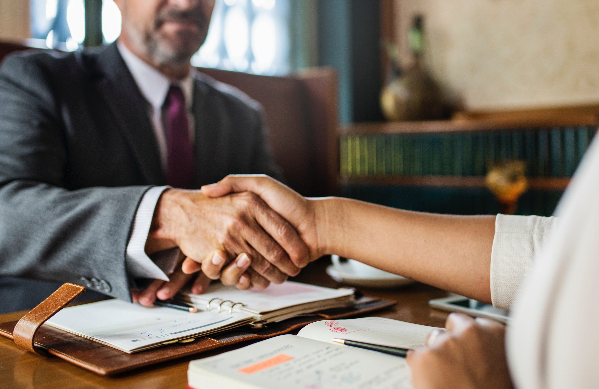 Two people are shaking hands across a desk with open notebooks and paperwork, suggesting a business meeting or agreement—perhaps an acquisition discussion involving Advanced Office Automation—in a professional GoodSuite office environment.