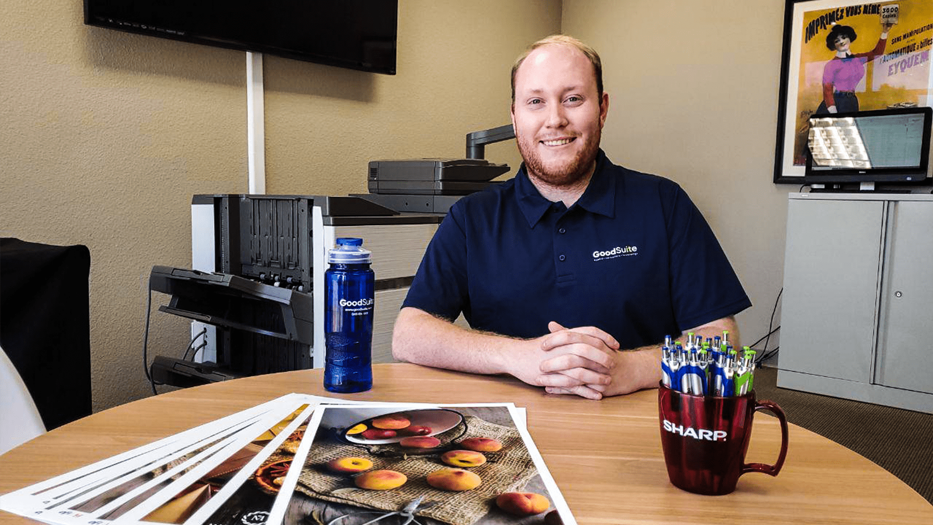 A smiling Summer Intern in a navy polo shirt sits at a round office table with printed photos, a blue water bottle, and a mug filled with pens. Office equipment and a framed poster are in the background.