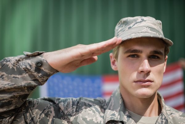 A young man in military uniform salutes while standing in front of a blurred American flag backdrop, honoring Military Appreciation Day.