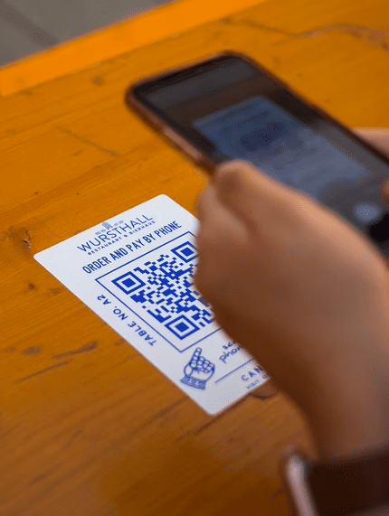 A person demonstrates the effective use of QR codes by holding a smartphone over a code printed on a WURSTHALL card to order and pay, showcasing how print materials can streamline dining at a wooden table.