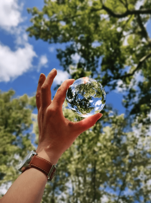 A hand wearing a brown watch holds up a clear glass sphere that reflects trees and the blue sky, emphasizing the beauty of the environment against a blurred background of leafy green trees.