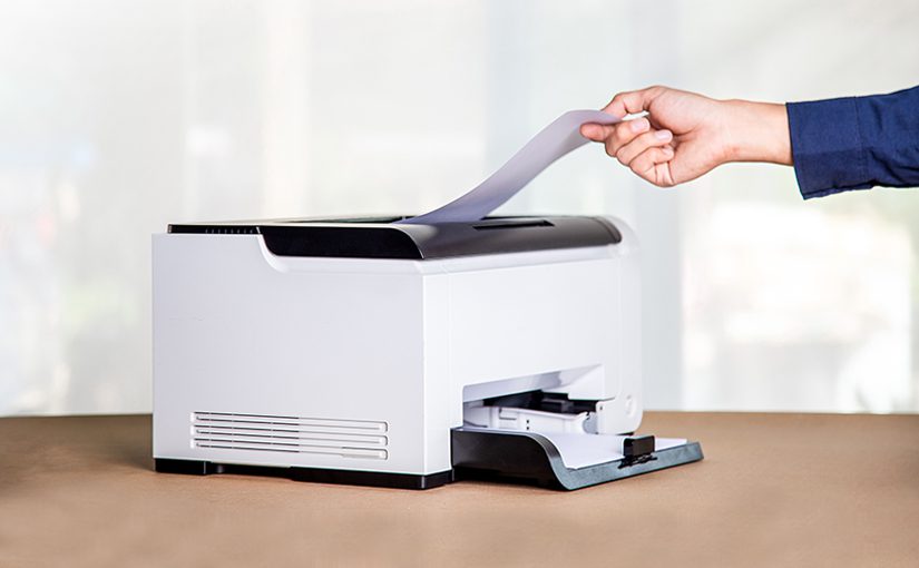 A person’s hand is placing a sheet of paper into a white office printer on a brown surface, with a blurred light background.