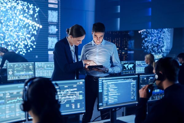 People in a modern control room work at computer stations with multiple screens displaying data and graphs. Two individuals in the center discuss information using a tablet, highlighting the IT benefits of Managed IT Services. Digital maps fill the background displays.