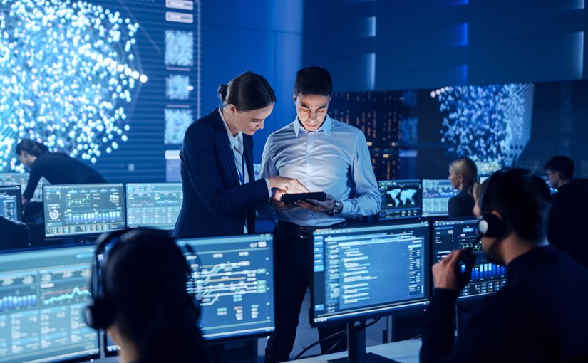People in a modern control room work at computer stations with multiple screens displaying data and graphs. Two individuals in the center discuss information using a tablet, highlighting the IT benefits of Managed IT Services. Digital maps fill the background displays.