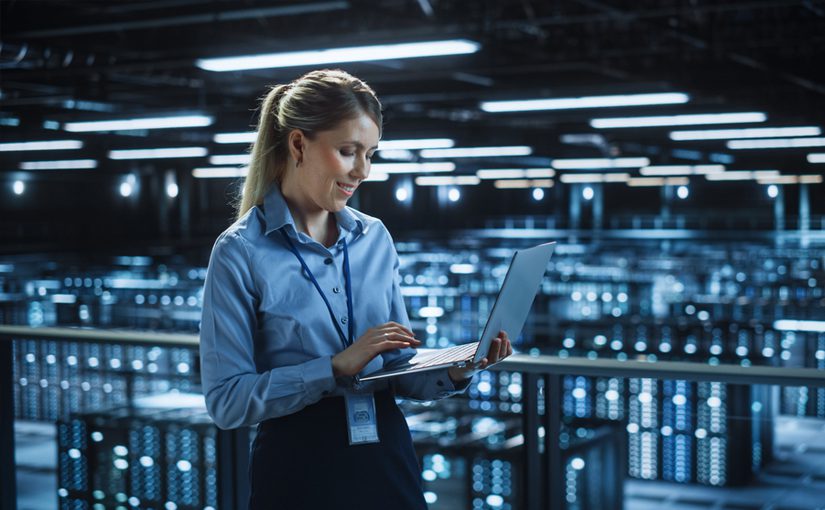 A woman in business attire stands in a modern data center, smiling while using a laptop. Rows of illuminated server racks fill the large, high-tech room—highlighting the efficiency of Managed IT Services in 2022.