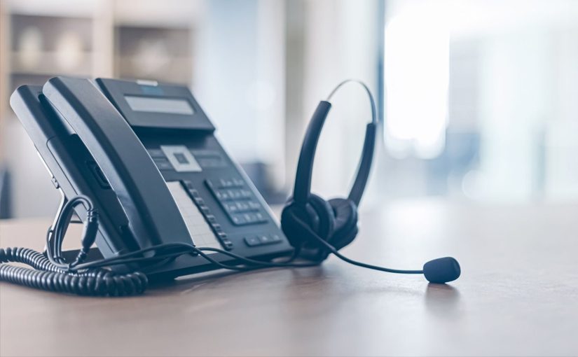A black office desk phone with a corded handset sits on a wooden desk next to a black headset with a microphone, showcasing modern VoIP systems in a bright office setting.