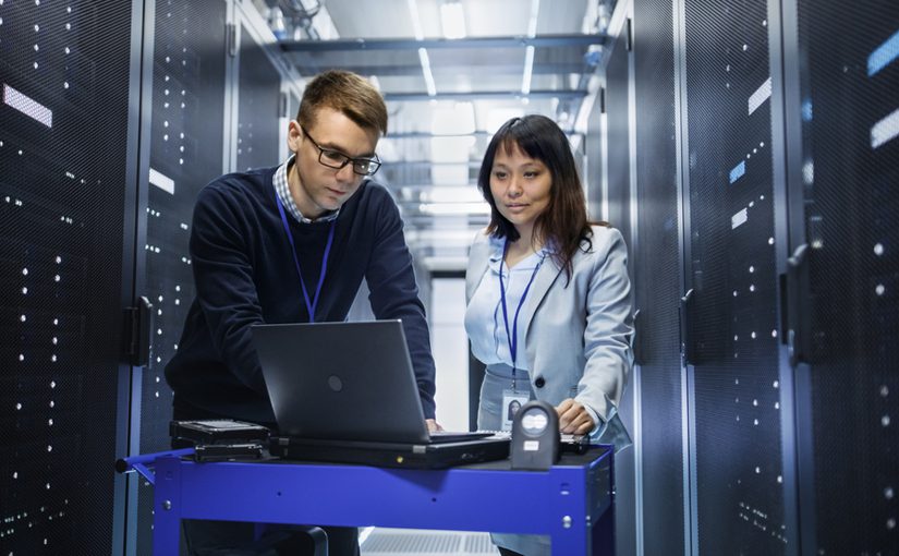 Two IT professionals, a man and a woman, stand in a server room looking at a laptop on a cart, delivering Managed IT Services. They wear ID badges and are surrounded by tall server racks with visible cables and lights.
