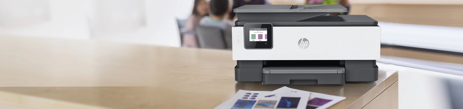 An HP printer, featured among the Best Printers 2023, sits on a wooden desk with colorful documents in front. In the blurred background, people are seated at a table, suggesting an office or collaborative workspace.