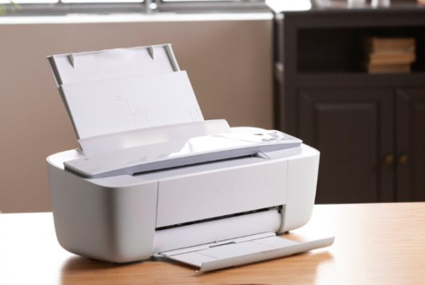 A white office printer sits on a wooden desk in a home office setting, with paper loaded in the tray. A plant and a cabinet with books and decor are visible in the softly lit background—ideal for those seeking the best printers for offices.