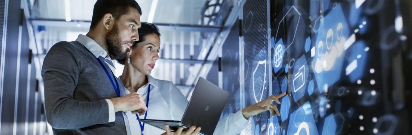 Two professionals stand in a server room, analyzing data icons on a large transparent digital screen. One holds a laptop while the other points at the display, illustrating collaboration in Managed IT Services.