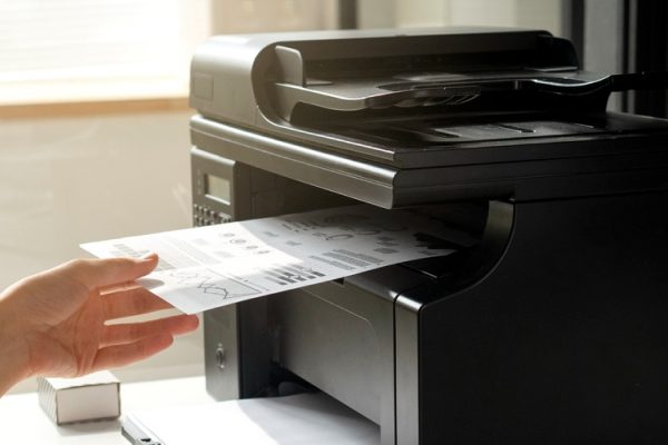 A person loads a document into a black printer or copier in an office, highlighting the benefits of Managed Print Services for small businesses, with a computer monitor displaying file folders and a progress bar visible nearby.
