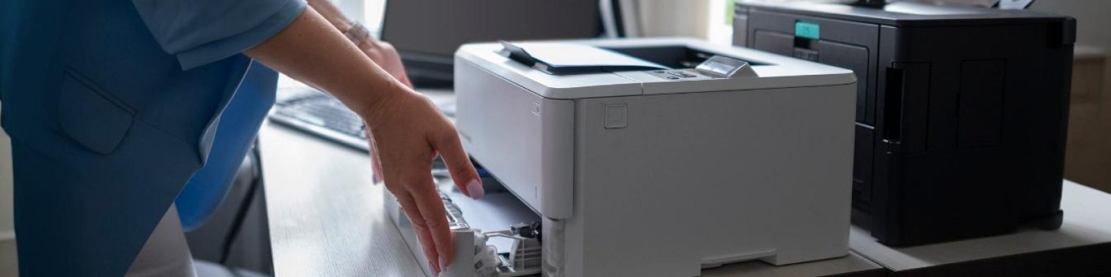 A person stands at a desk, loading paper into a white printer—ideal for small businesses—with another black printer and a computer monitor nearby, showcasing options among the top 10 commercial printers.