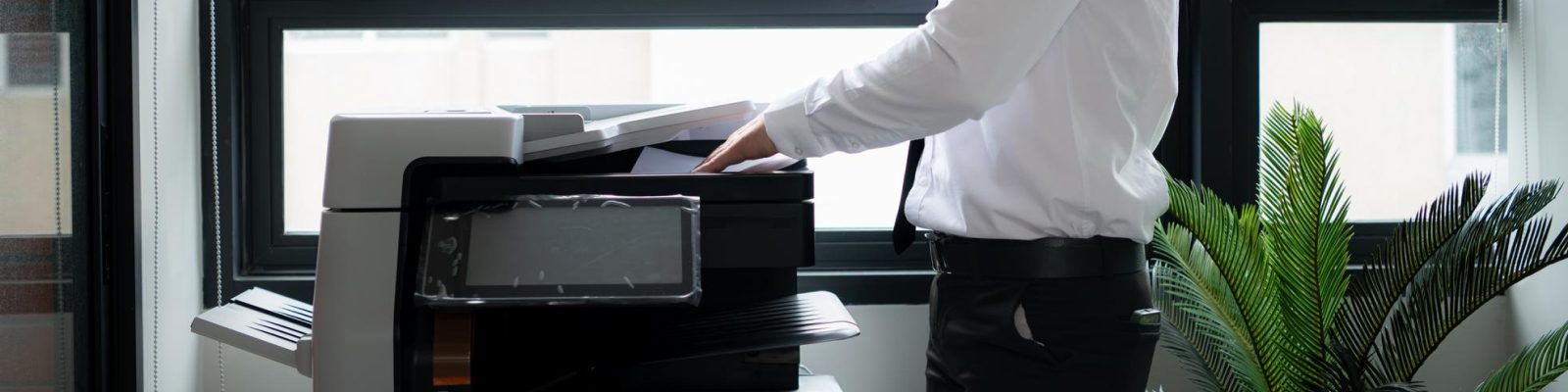 A person in business attire stands next to a copier, showcasing Print Solutions in a brightly lit office with large windows and a potted plant—highlighting the benefits these services offer construction companies.