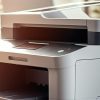 A modern office printer sits on a desk in a bright, sunlit workspace—ideal for law firms—with shelves and plants in the background.