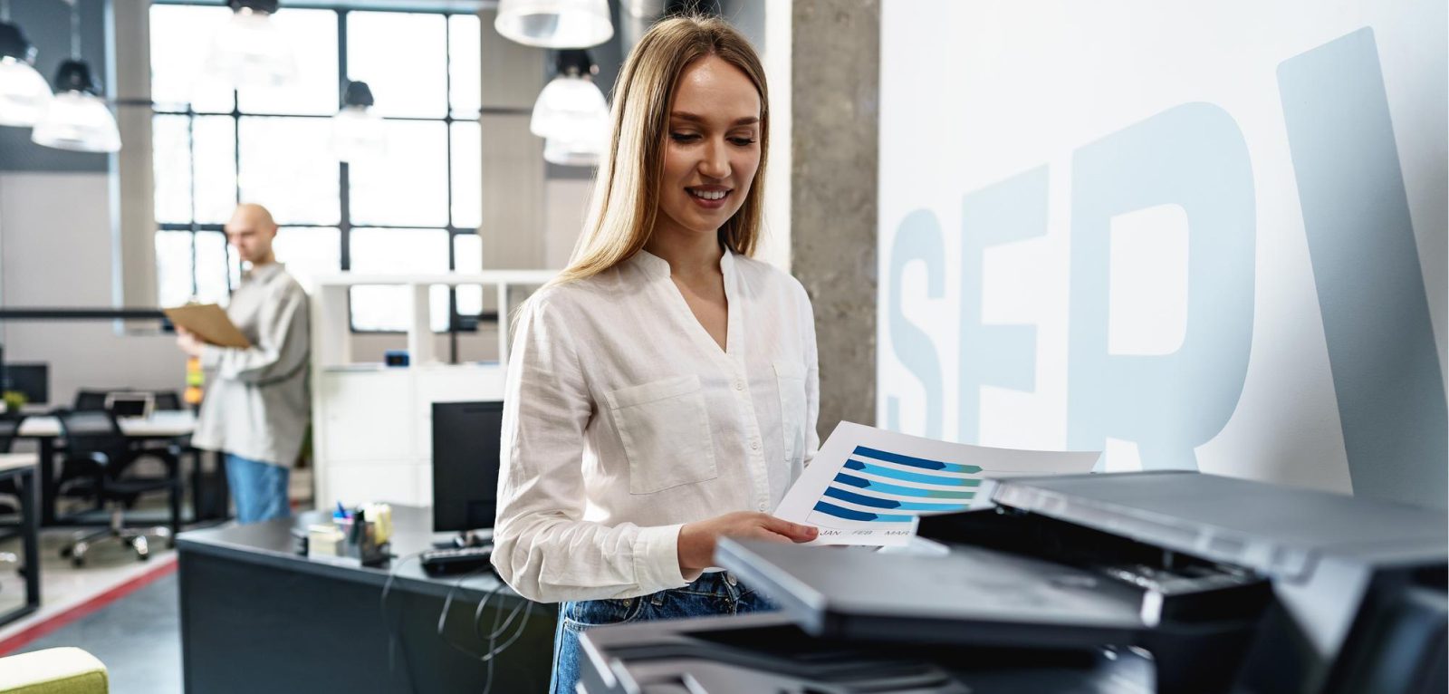 A woman stands by an office printer, holding printed charts and smiling. In the background, a man with a clipboard works at a desk in a modern, well-lit office—reflecting how Managed Print Services help SMBs reduce costs efficiently.