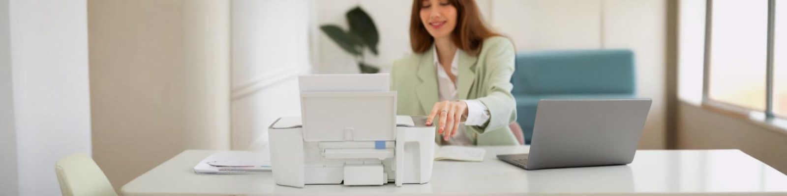 A woman in a light green blazer sits at a desk with a laptop, operating a white printer. The bright, minimal workspace suggests she might be following a secure print management guide to ensure print security and efficiency.