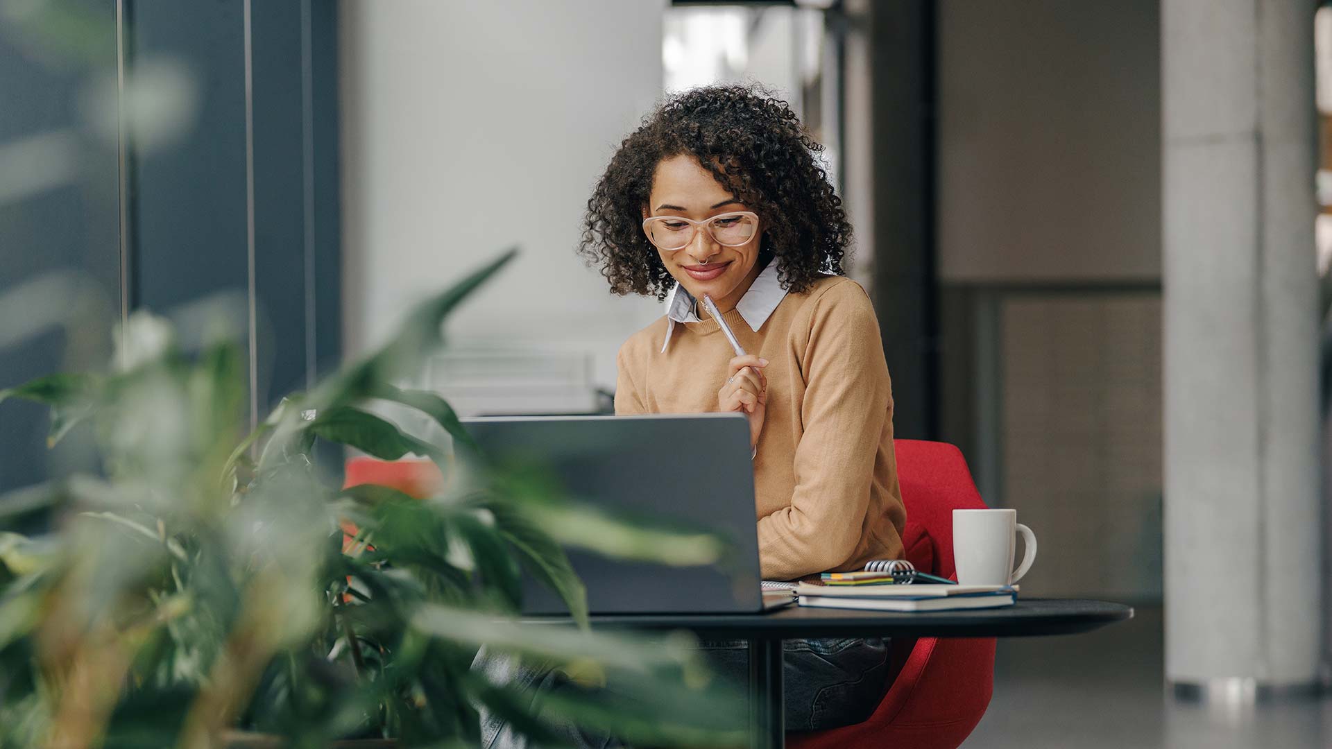 A woman with curly hair and glasses sits at a table, smiling at her laptop. She holds a pen to her chin, with a notebook and coffee mug next to her. Green plants are visible in the foreground.