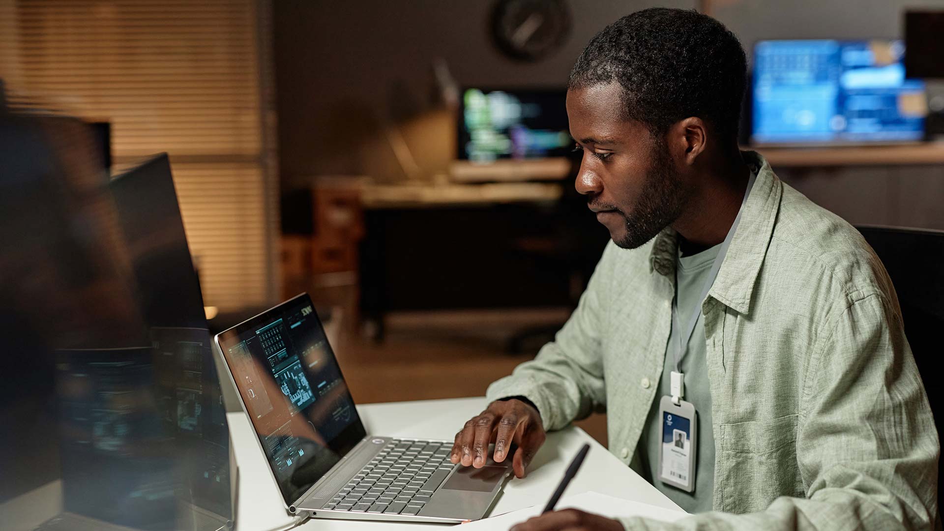 A man wearing a security badge sits at a desk, focused on an IT Support dashboard on his laptop displaying security data, in a dimly lit office with computer monitors in the background.