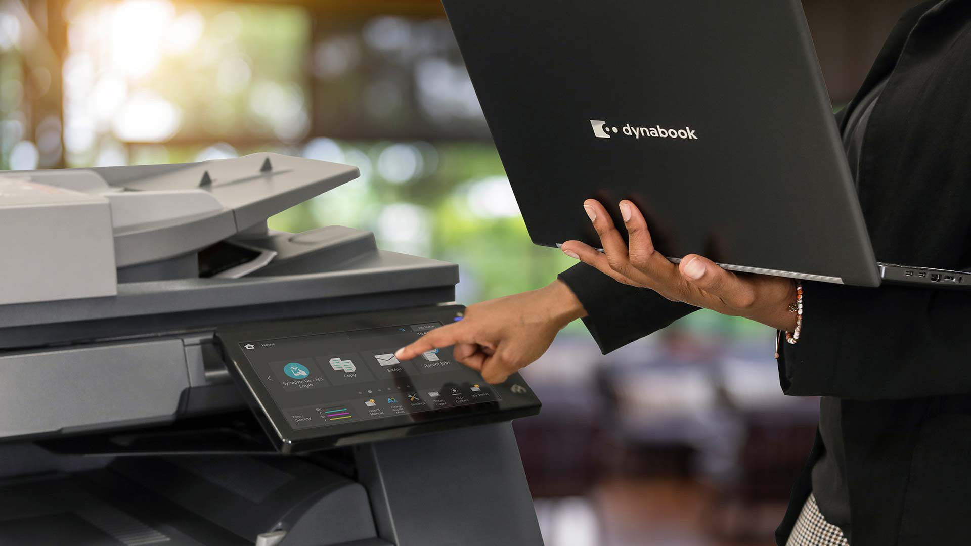 A person holding a Dynabook laptop uses the touchscreen control panel of a modern printer in an office setting with blurred background.